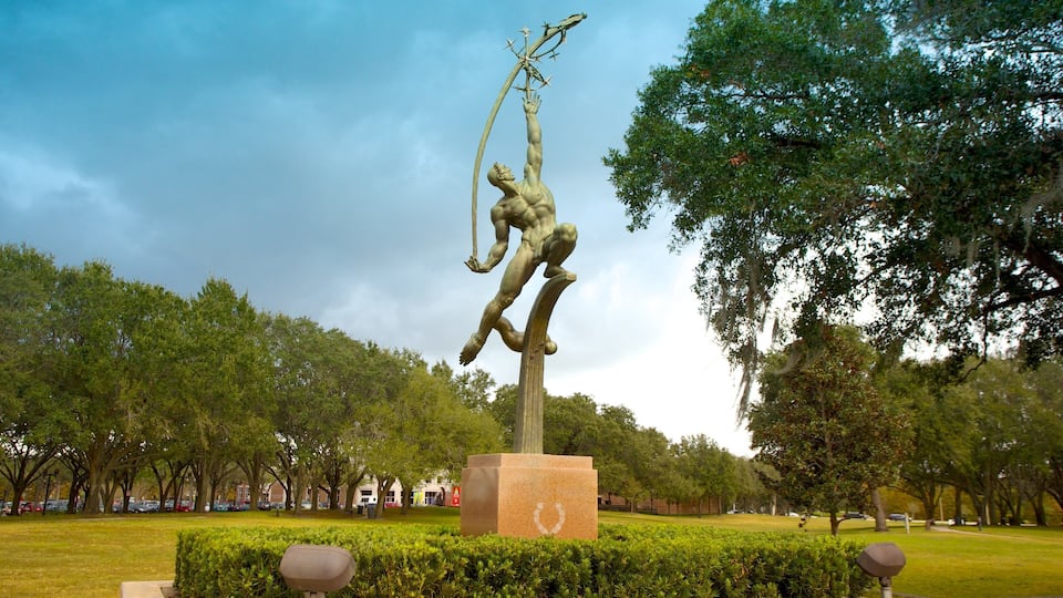 A bronze statue of a muscular man holding a spear in a park with trees and a clear sky.