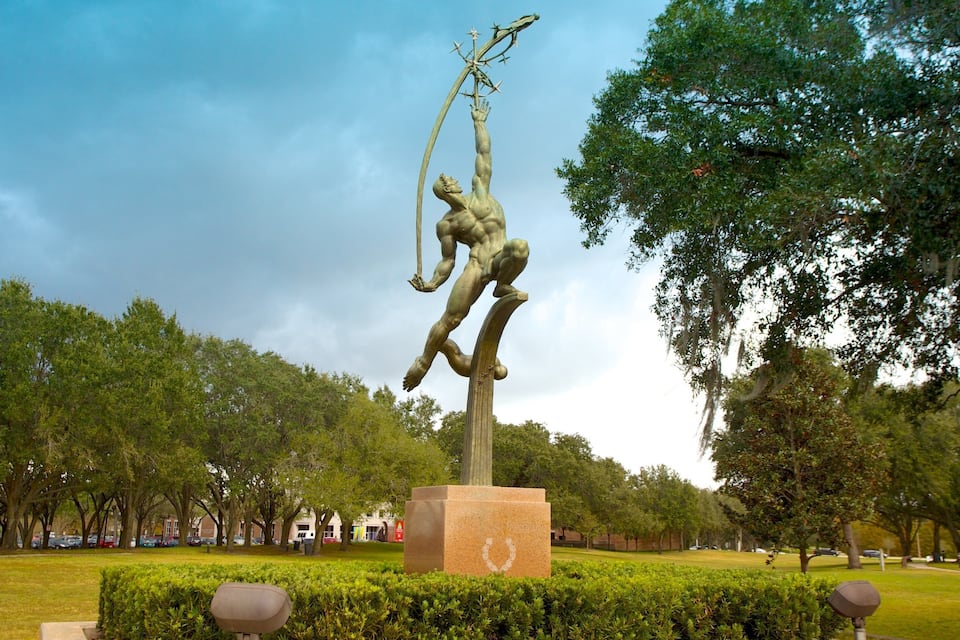 Une statue de bronze représentant un homme musclé tenant une lance, dans un parc arboré, sous un ciel dégagé.