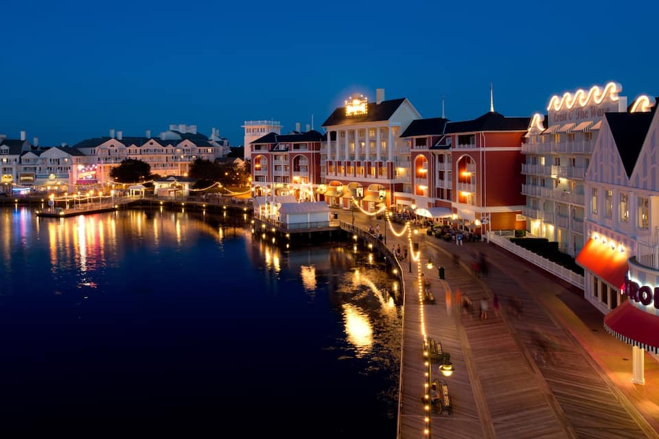 A waterfront promenade with illuminated buildings and a boardwalk reflecting on the water.