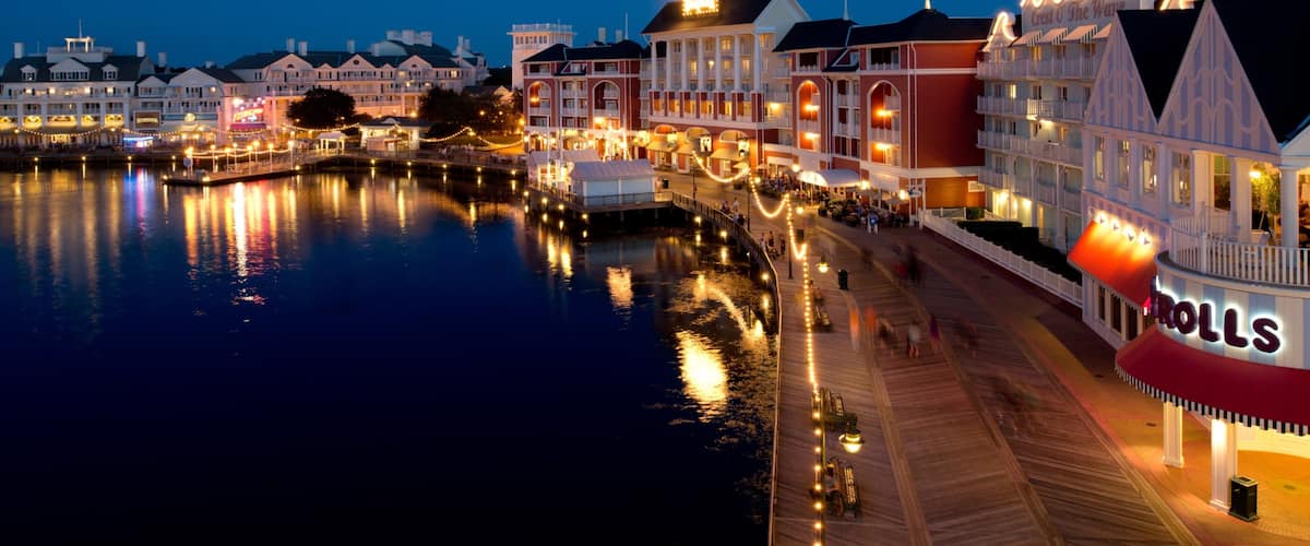 A waterfront promenade with illuminated buildings and a boardwalk reflecting on the water.