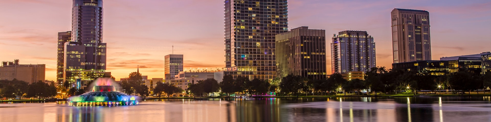 Silhueta urbana ao entardecer, com edifícios iluminados e um lago tranquilo refletindo as luzes.