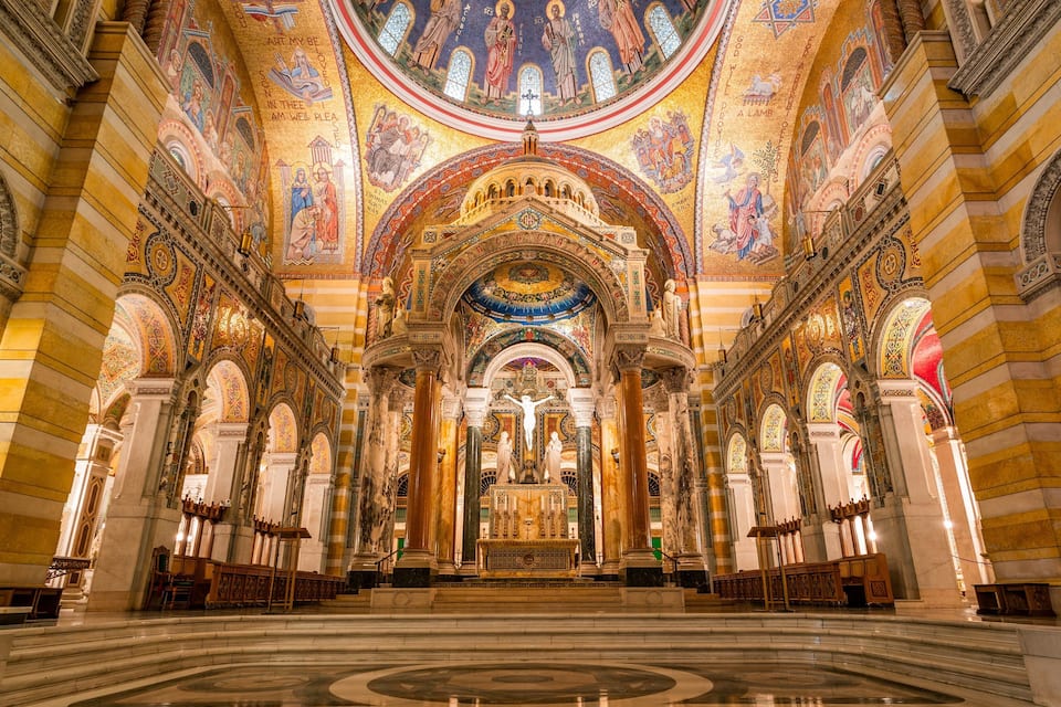 Interior imponente de uma catedral com mosaicos elaborados, um grande altar central e uma janela circular de vitrais.