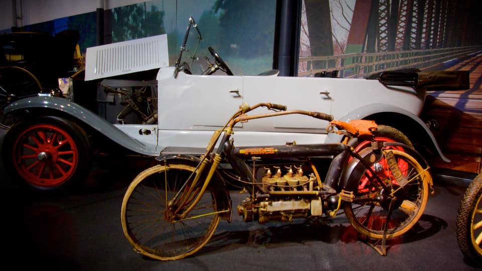 A vintage car with a bicycle engine attached, displayed indoors with a mural in the background.
