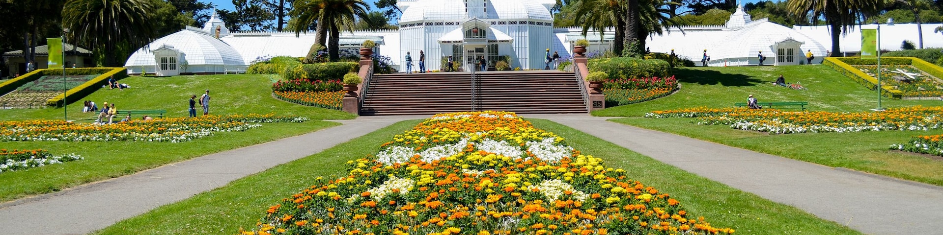 Um jardim botânico com uma estrutura central em forma de cúpula branca, rodeado por canteiros de flores bem cuidados e palmeiras.