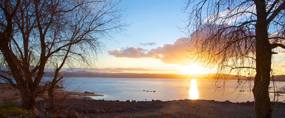 A sunset over a calm lake with rocky shores and leafless trees.
