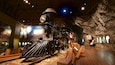 A steam locomotive in a museum exhibit with a rocky backdrop and a sign indicating a historical achievement.