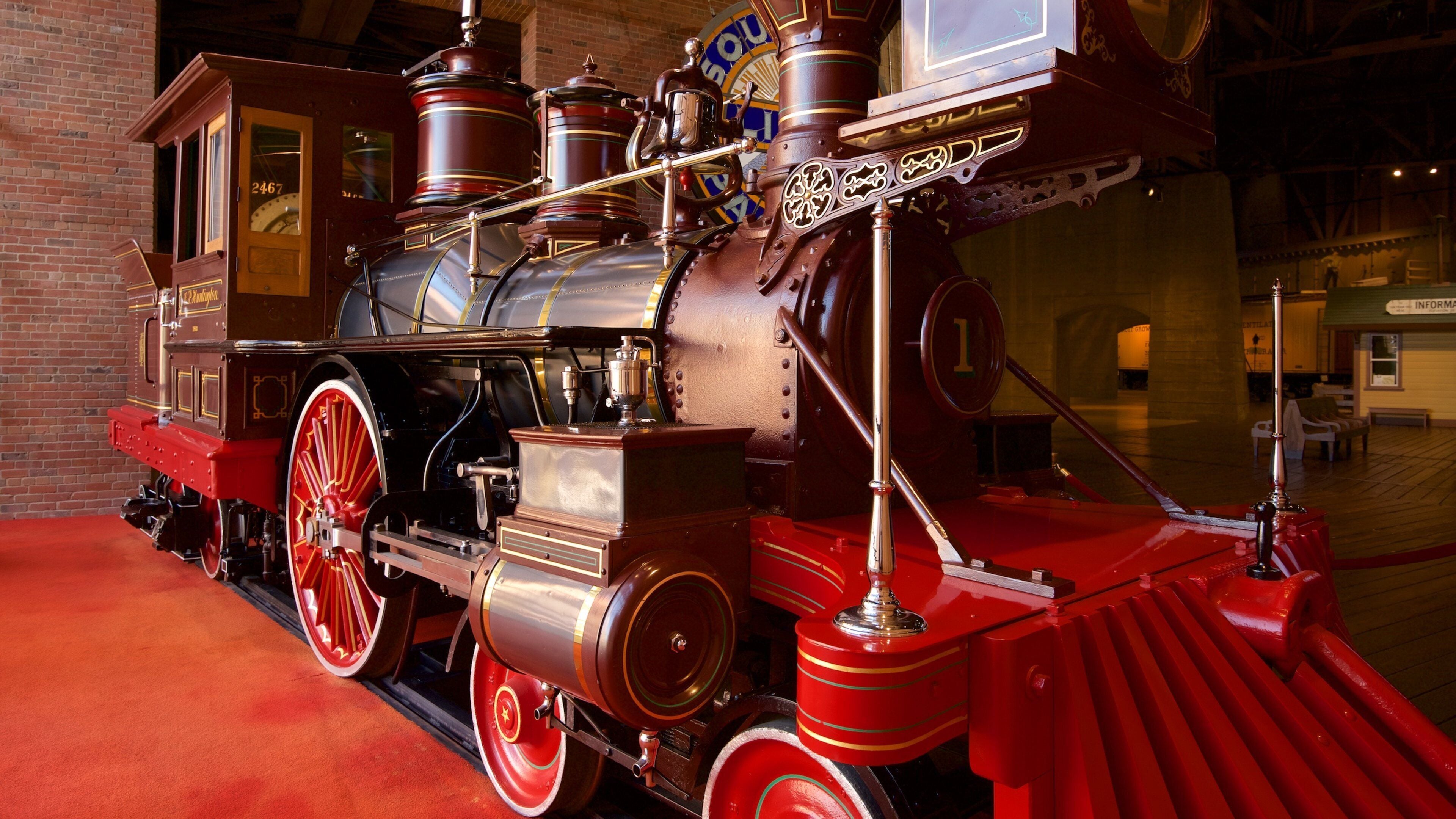 A vintage steam locomotive with red wheels and brass fittings, displayed indoors with a brick wall background.