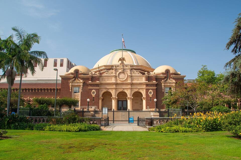 A historic building with a dome and an eagle statue, surrounded by well-maintained gardens and palm trees.