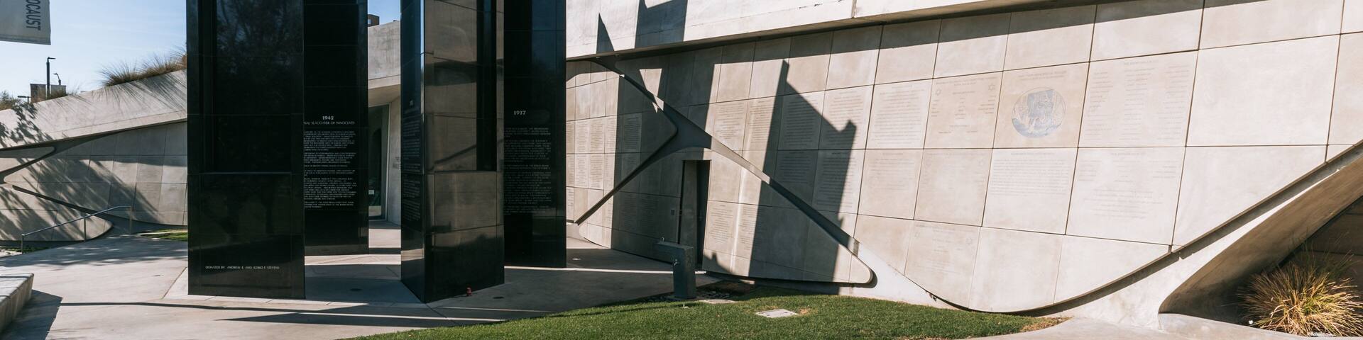 Un monumento con paneles negros altos y grabados, frente a un muro de concreto y un área verde.