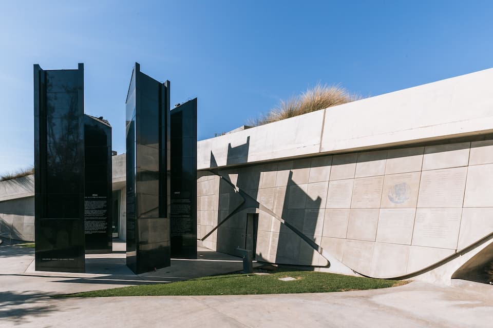 A memorial with tall black panels and inscriptions, set against a concrete wall with a grassy area in front.