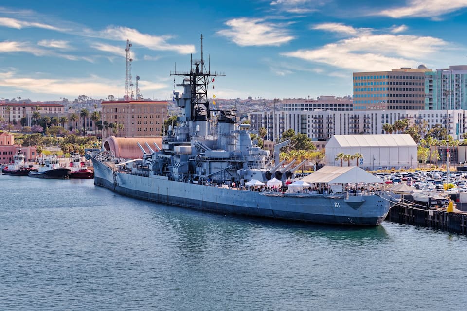 A large naval ship docked at a port with a cityscape in the background.