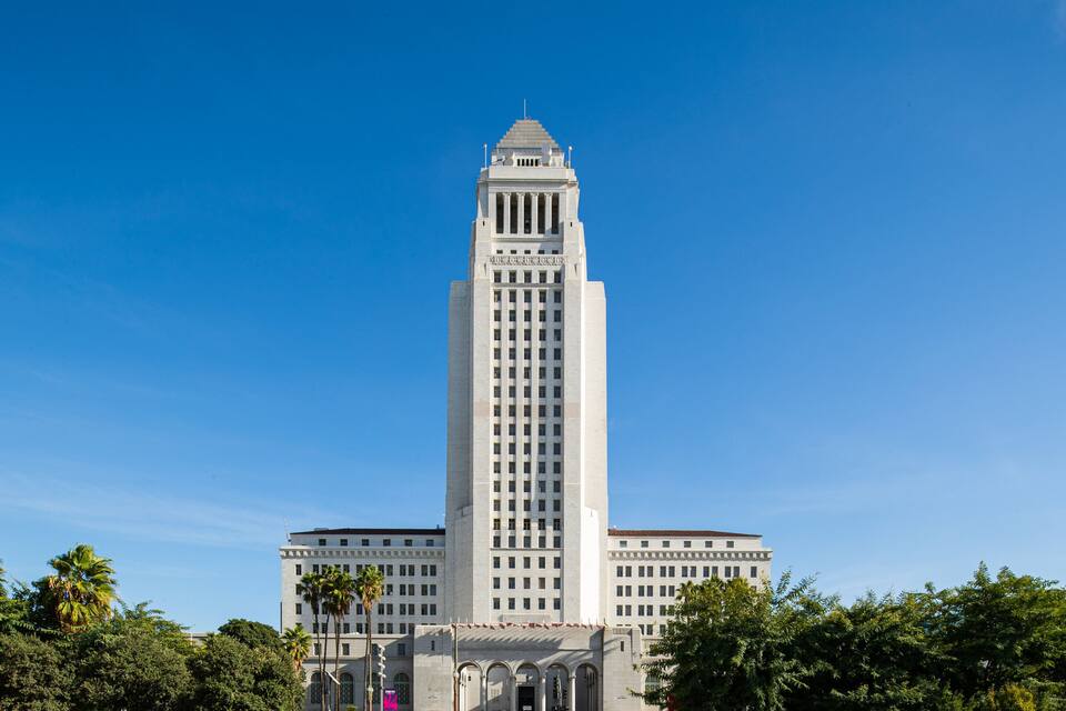 A tall, white building with a triangular roof and columns, surrounded by trees and a clear blue sky.