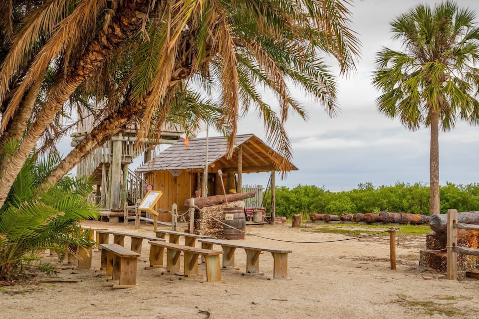 A rustic wooden structure with benches, surrounded by palm trees and a sandy area.