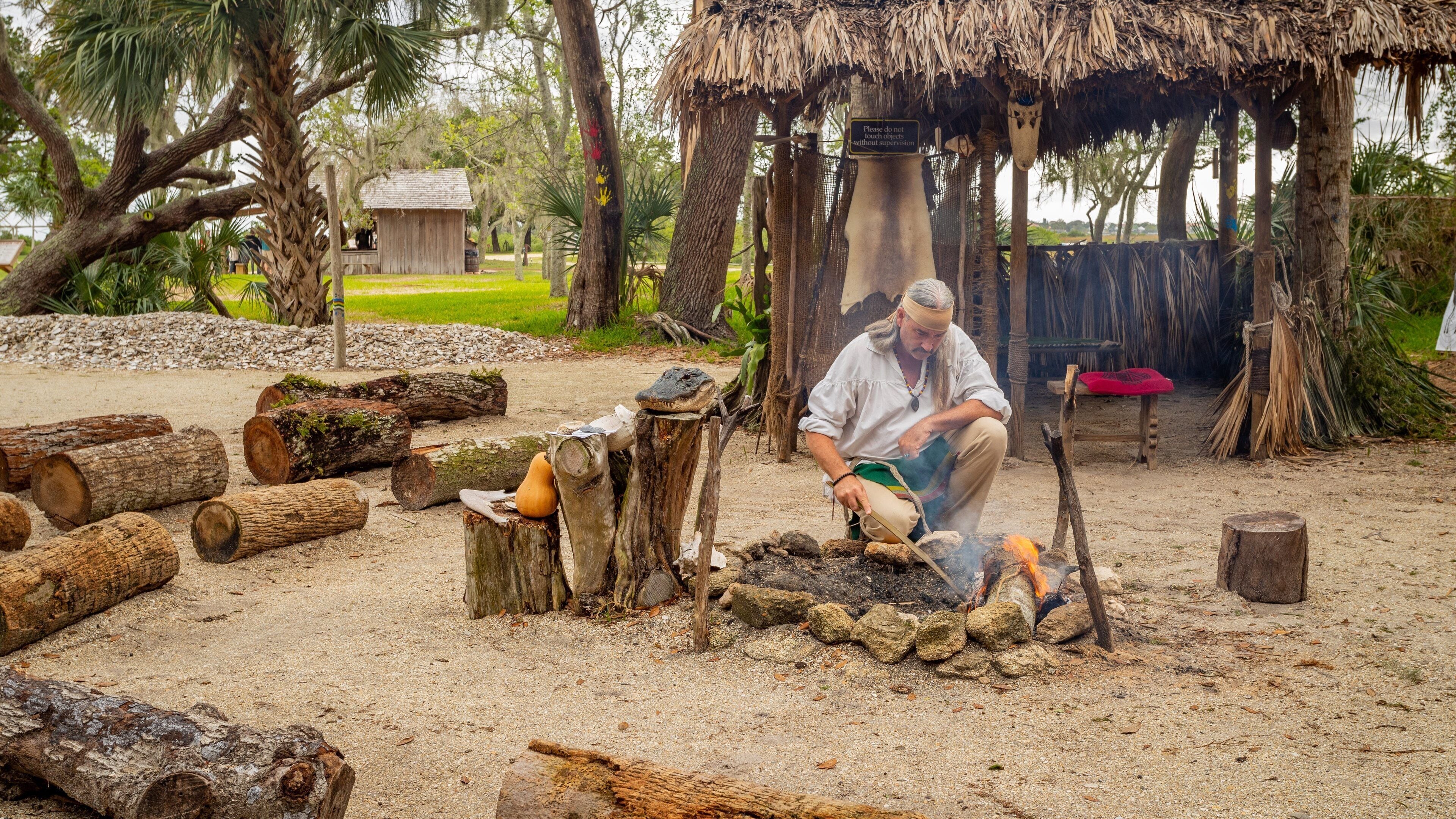 A man cooking outdoors with a fire pit, surrounded by logs and a thatched-roof structure.