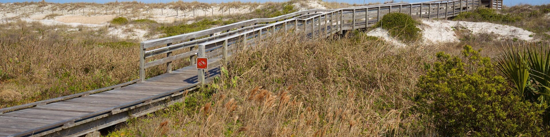 Un muelle de madera que lleva a una playa con dunas y vegetación escasa.