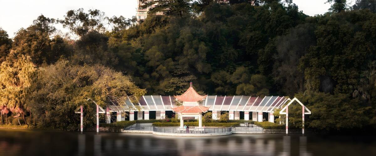 A traditional pavilion with a tiled roof, surrounded by lush greenery and a calm lake.