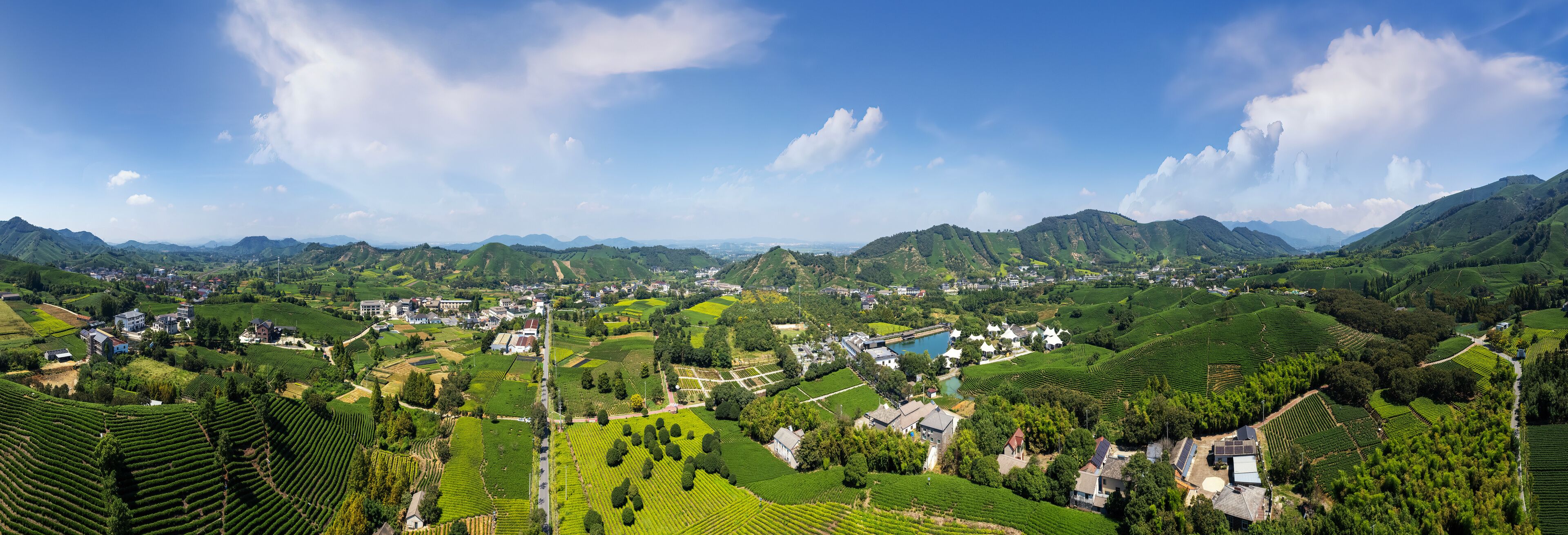 A panoramic view of a lush valley with terraced fields, a small village, and distant mountains under a clear sky.