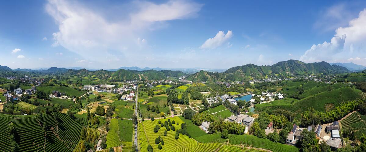 A panoramic view of a lush valley with terraced fields, a small village, and distant mountains under a clear sky.