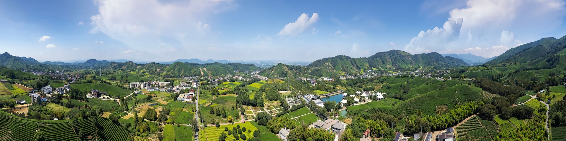 A panoramic view of a lush valley with terraced fields, a small village, and distant mountains under a clear sky.