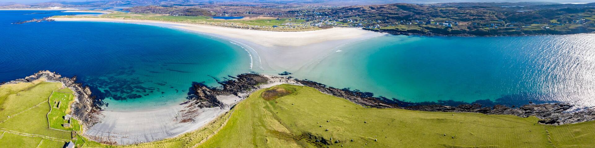 A coastal landscape with a sandy beach, clear blue waters, and green fields.