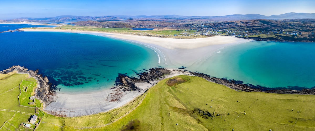 A coastal landscape with a sandy beach, clear blue waters, and green fields.