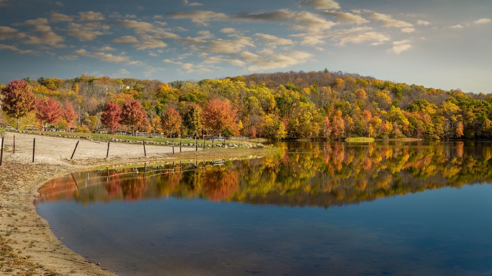 A calm lake reflecting autumn trees, a sandy shore, and a distant hill with a cabin.