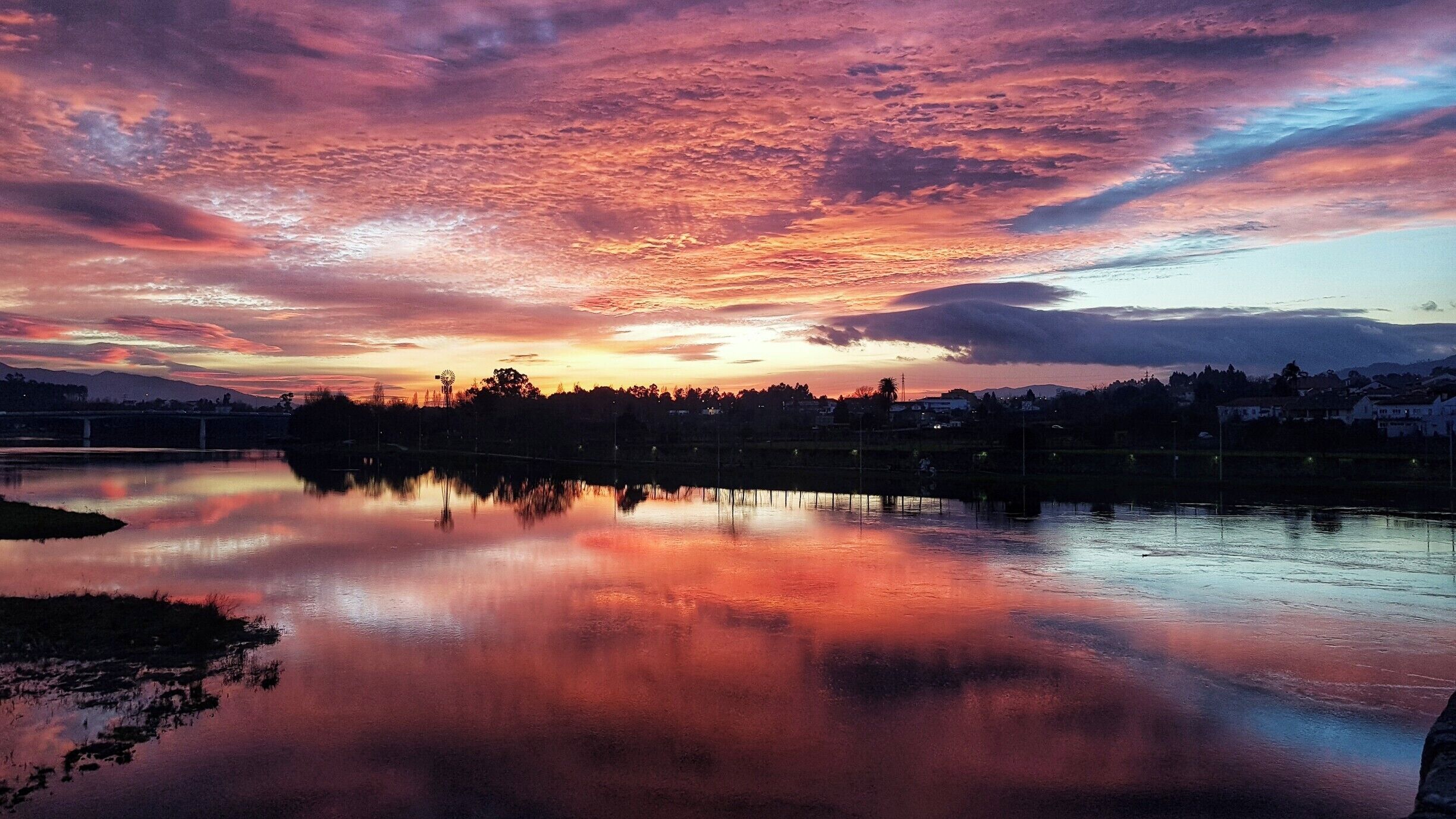 A sunset over a calm lake with a windmill in the foreground and a town in the background.