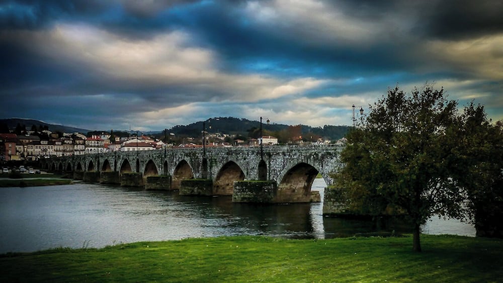 A stone bridge over a river with a grassy foreground and a cloudy sky.