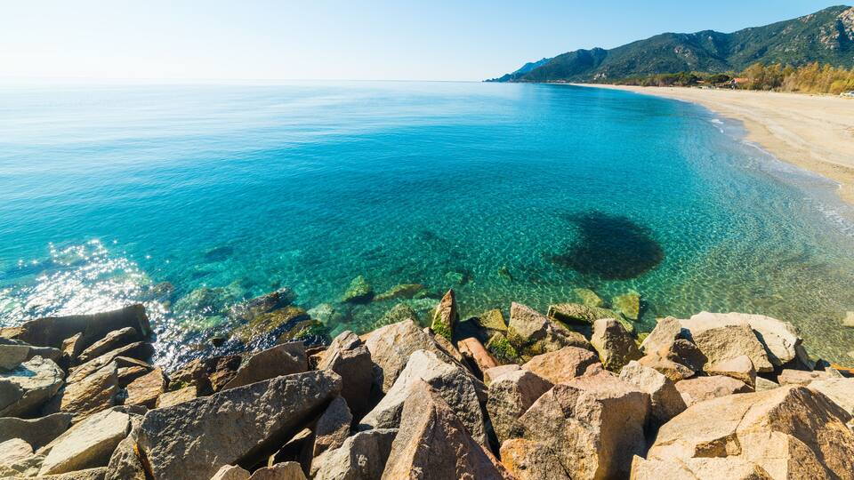 A clear blue sea, rocky shoreline, and a mountain in the distance.