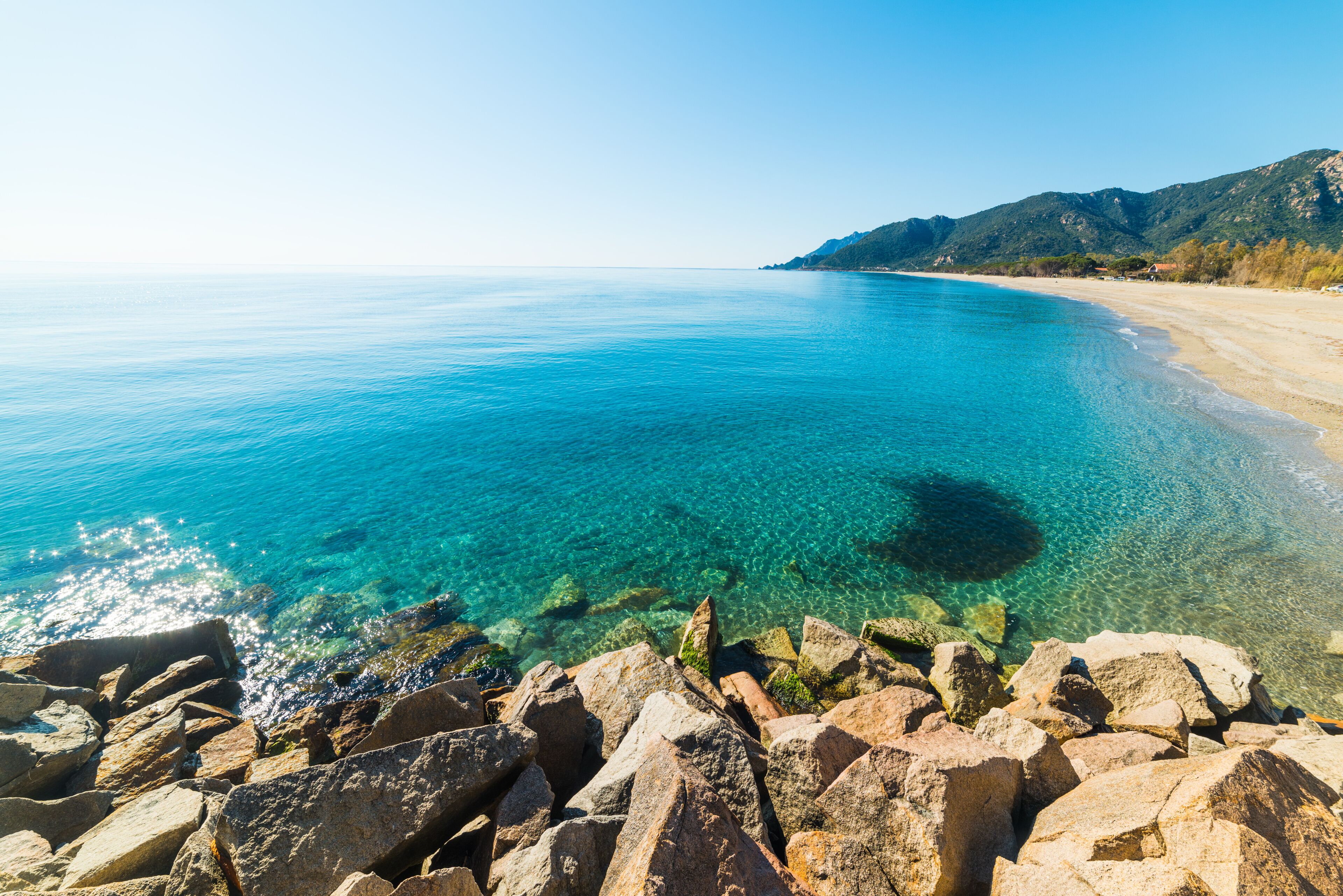 A clear blue sea, rocky shoreline, and a mountain in the distance.