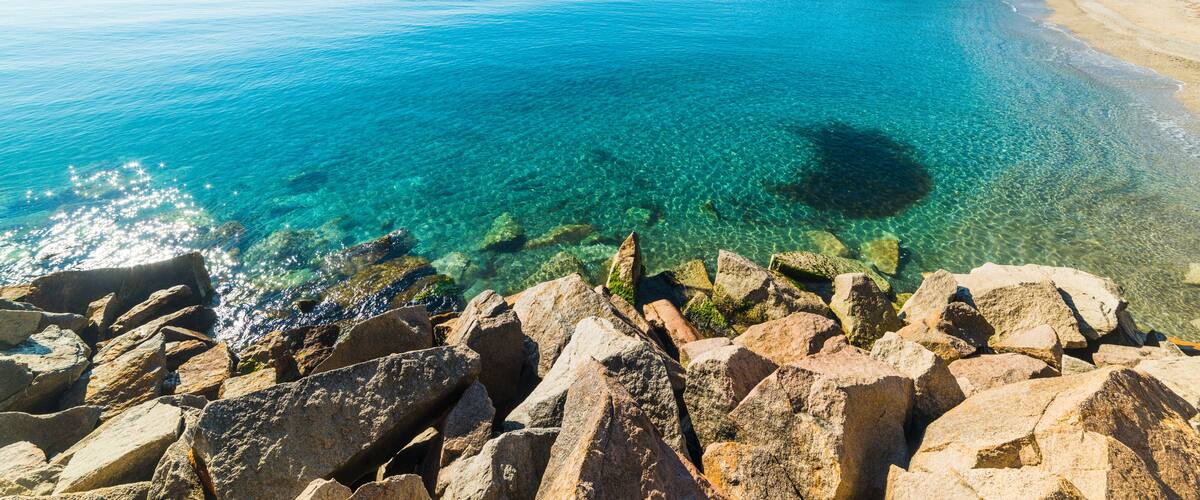 A clear blue sea, rocky shoreline, and a mountain in the distance.