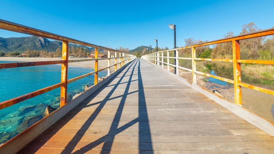 A wooden boardwalk with metal railings, leading to a body of water with clear blue water and a mountainous landscape in the background.