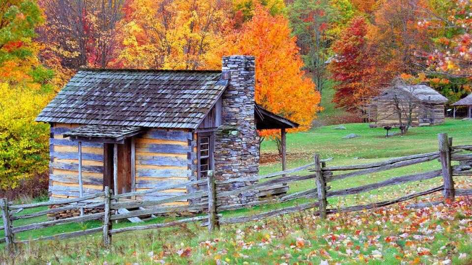 A rustic log cabin with a stone chimney, surrounded by a wooden fence and a vibrant autumn forest.