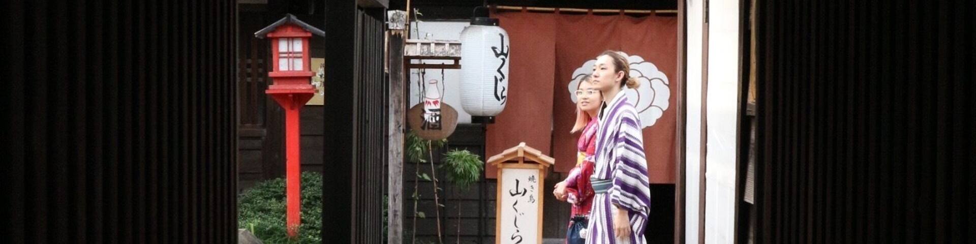 Un callejón tradicional japonés con edificios de madera, un farolillo rojo y una pareja vestida con atuendos tradicionales.