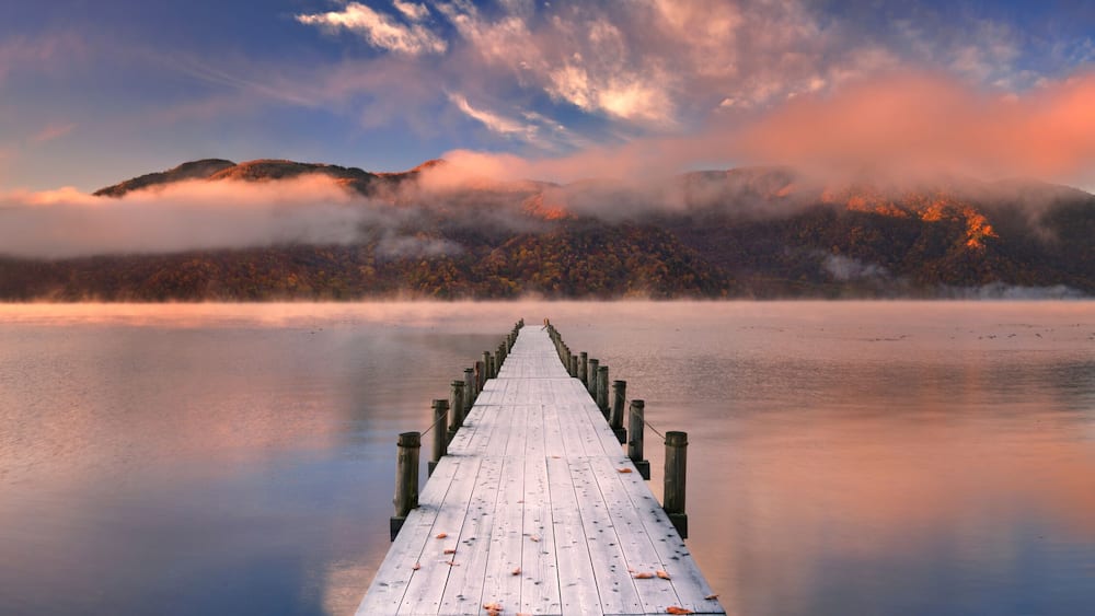 Eine Holzstege, der sich in ruhiges Wasser erstreckt, umgeben von Bergen und Herbstlaub.