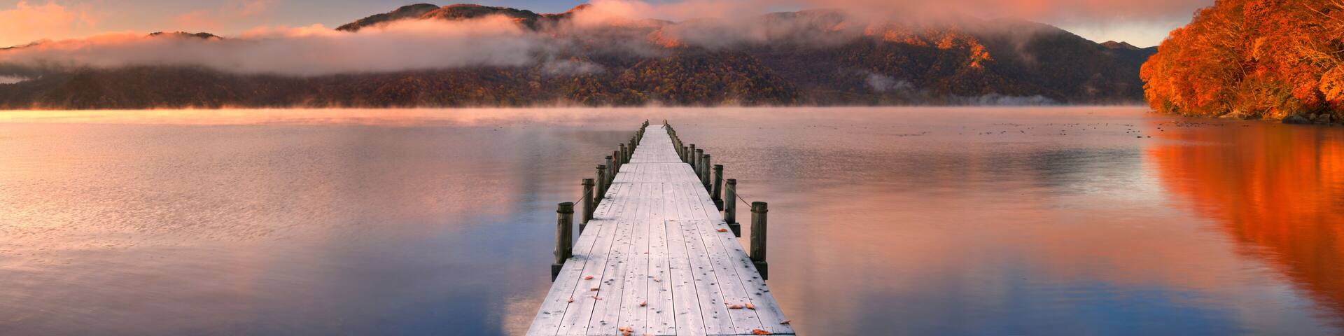 A wooden pier extending into calm waters, surrounded by mountains and autumn foliage.