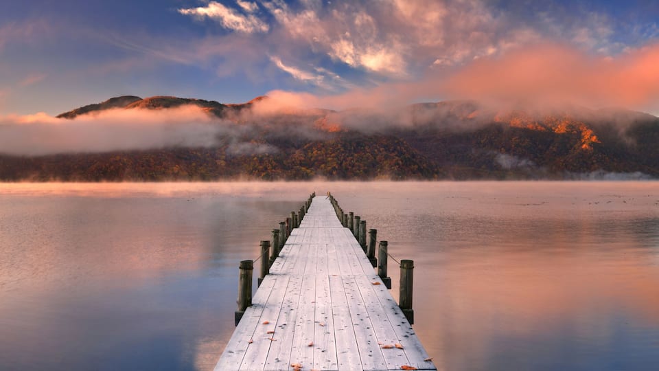 A wooden pier extending into calm waters, surrounded by mountains and autumn foliage.