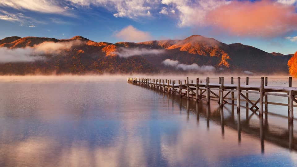 A calm lake with a wooden pier extending into the water, surrounded by mountains and autumn-colored trees.