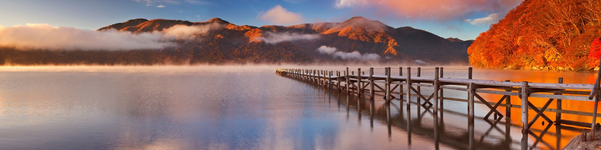A calm lake with a wooden pier extending into the water, surrounded by mountains and autumn-colored trees.