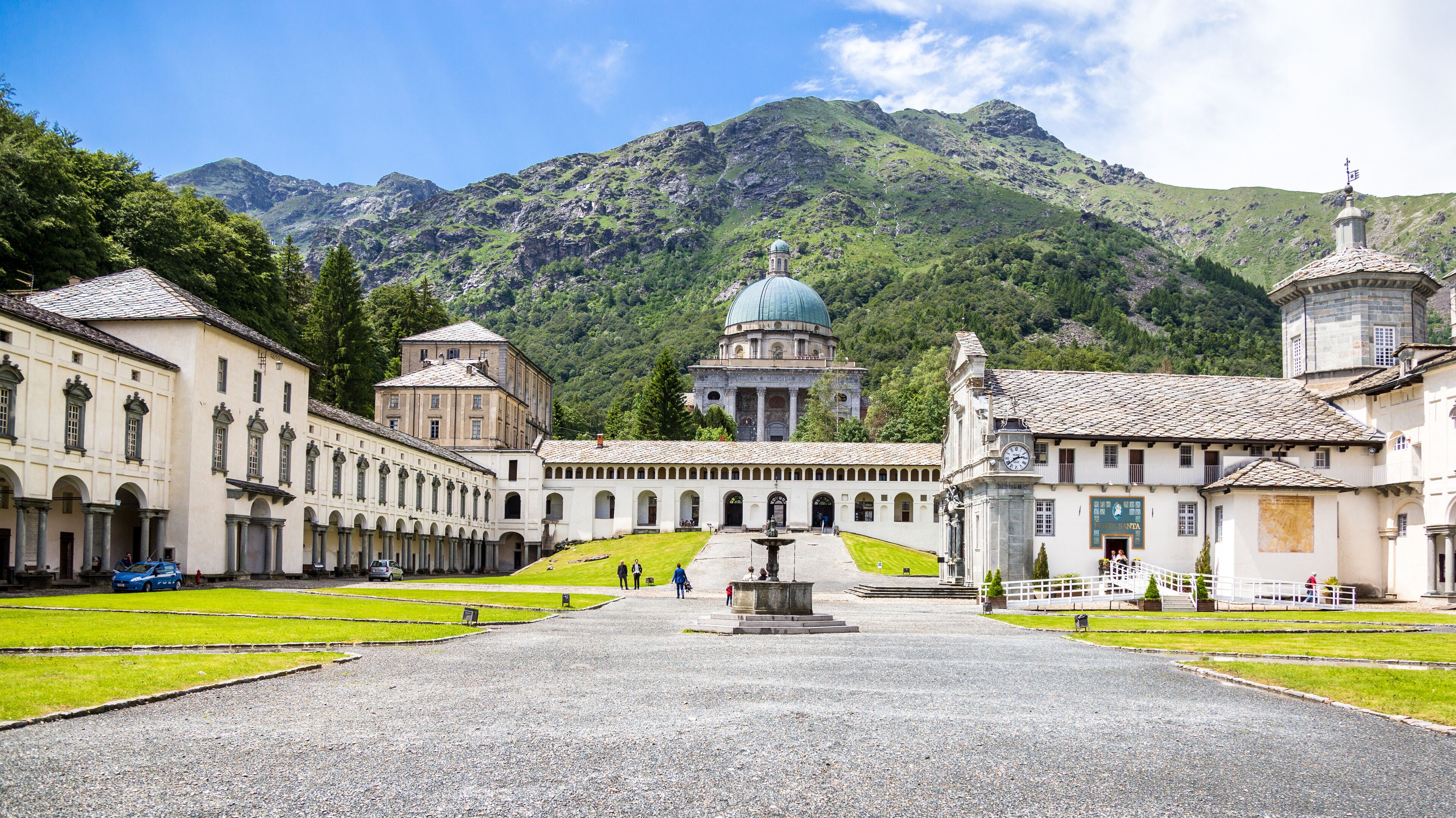 A historic courtyard with a central fountain, surrounded by classical architecture and a mountain backdrop.