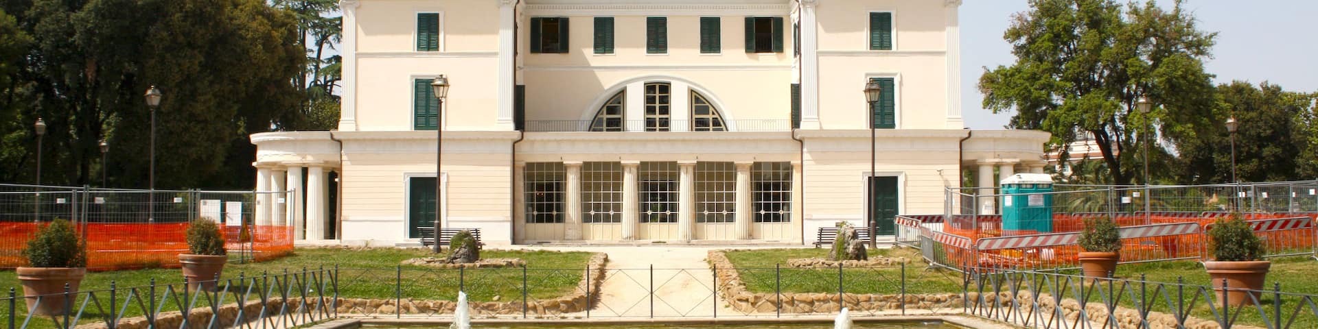 A large white building with a central arched entrance, surrounded by a formal garden with a fountain.