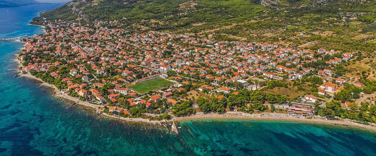A coastal town with red-roofed houses, a clear blue sea, and a mountain in the background.