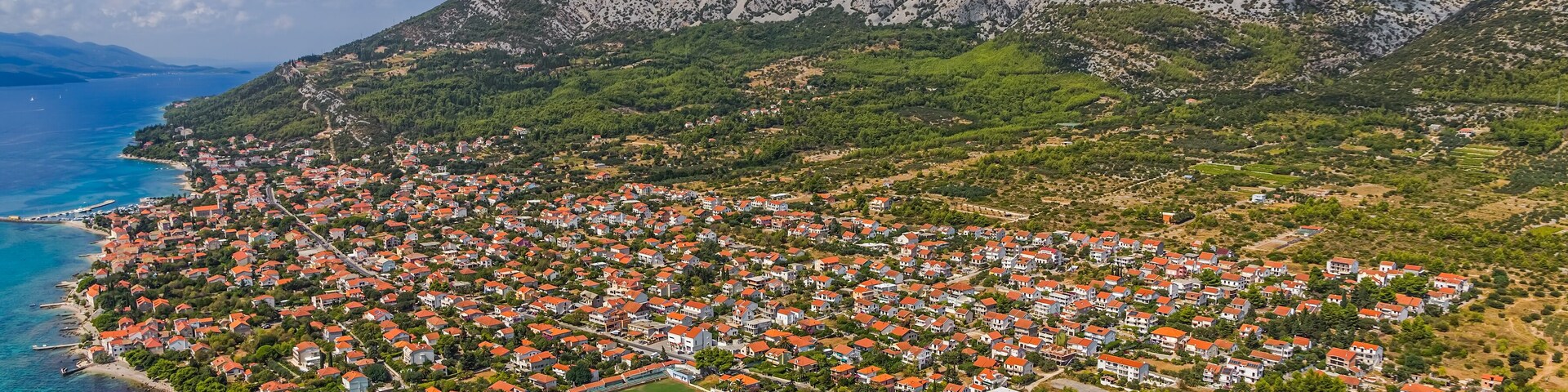 A coastal town with red-roofed houses, a clear blue sea, and a mountain in the background.