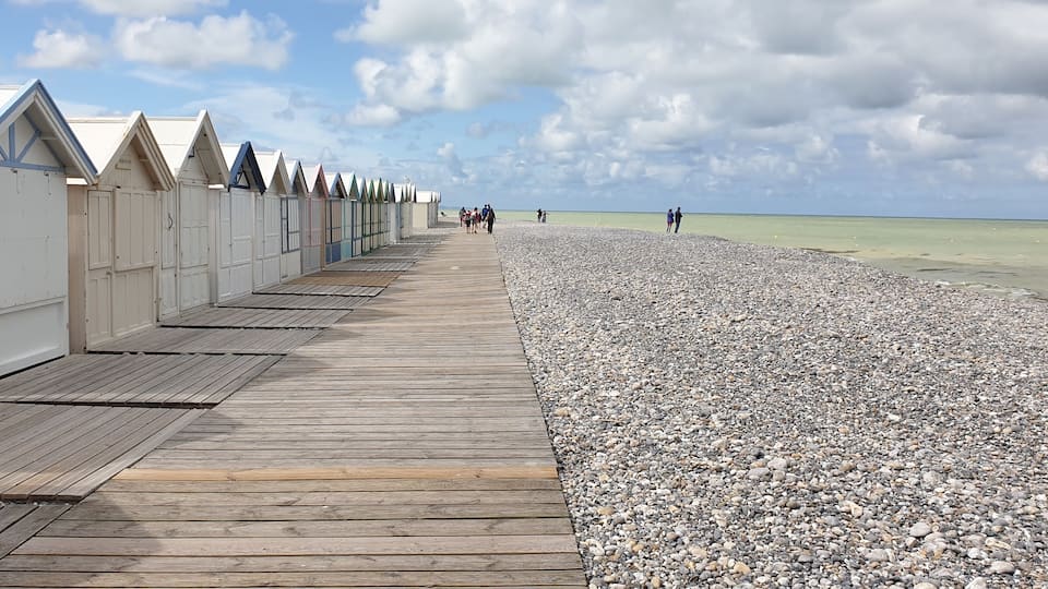 A wooden boardwalk leading to beach huts with a pebbled beach and a cloudy sky.
