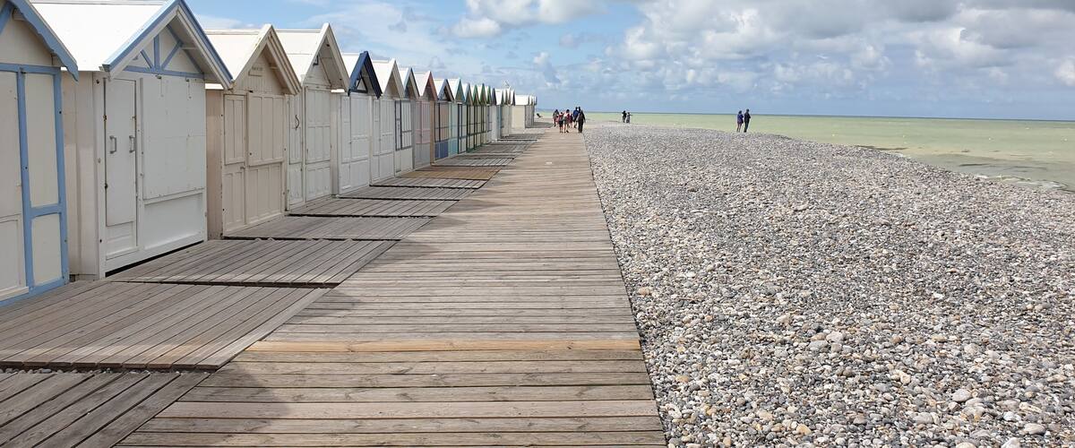 A wooden boardwalk leading to beach huts with a pebbled beach and a cloudy sky.