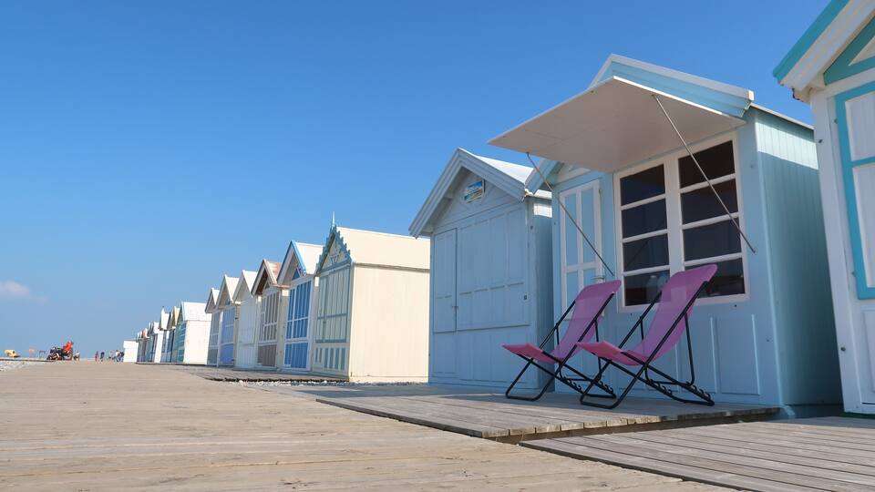 A row of beach huts with a wooden boardwalk and pink beach chairs in front.