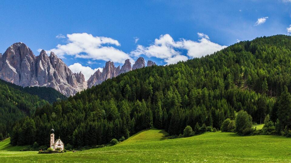 A green field with a church, forested hills, and rocky mountains.