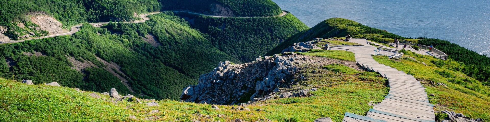 Un camino de montaña sinuoso, una pasarela de madera y vista al océano.