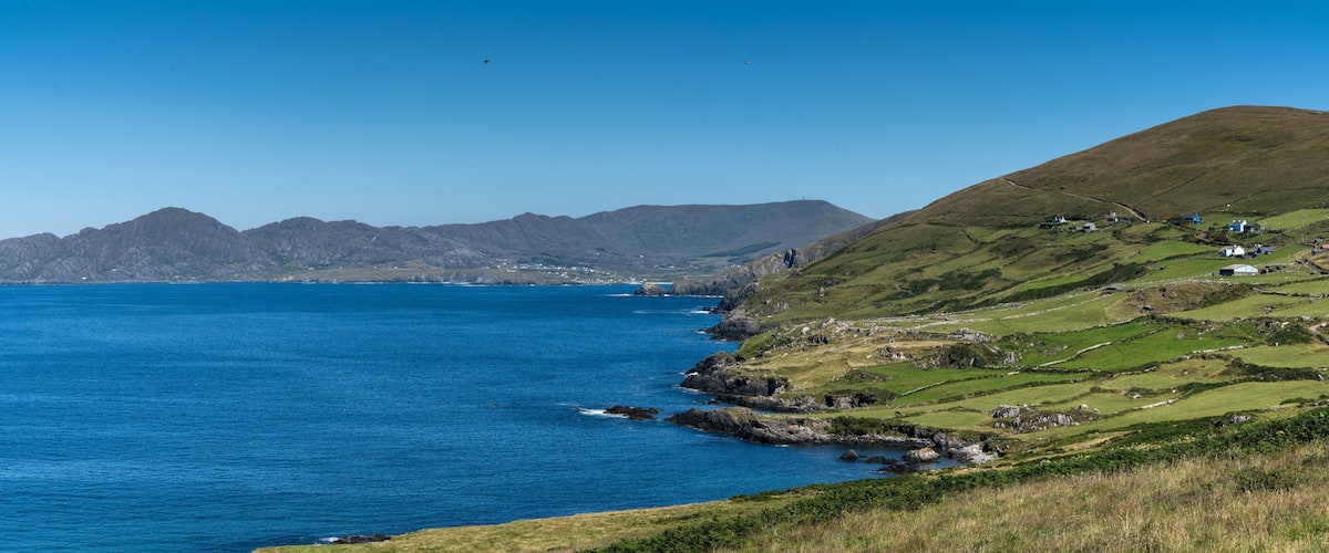 A coastal landscape with a clear blue sea, green hills, and scattered houses.