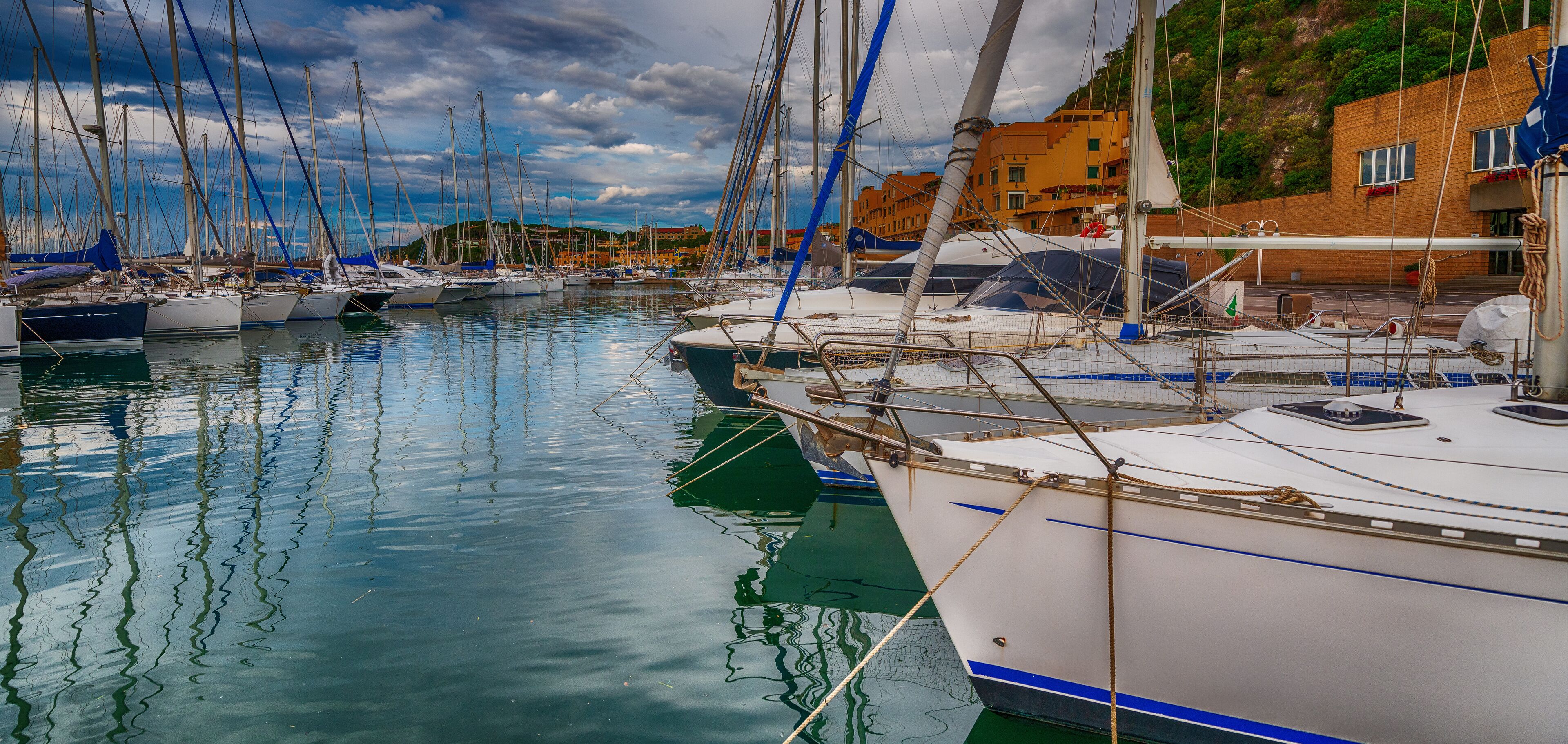 A marina with multiple sailboats docked, a building with red and yellow facades, and a calm water body reflecting the sky.